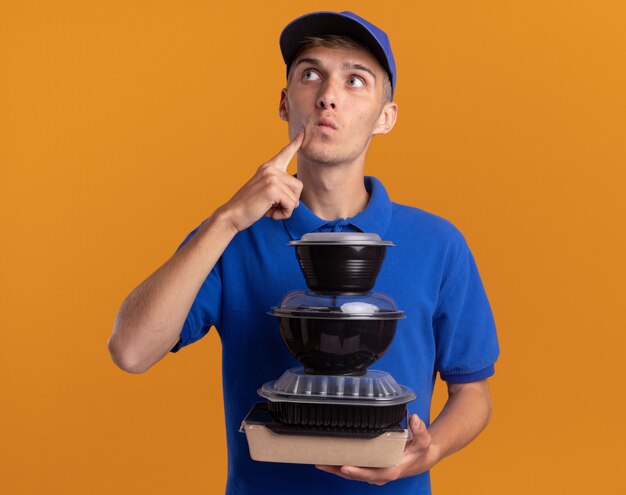 Thoughtful young blonde delivery boy puts finger on chin and holds food containers looking up isolated on orange wall with copy space