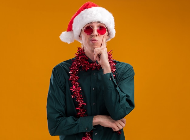 Thoughtful young blonde man wearing santa hat and glasses with tinsel garland around neck looking at camera keeping hand on chin isolated on orange background with copy space