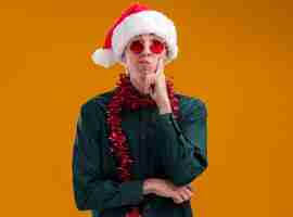 Free photo thoughtful young blonde man wearing santa hat and glasses with tinsel garland around neck looking at camera keeping hand on chin isolated on orange background with copy space