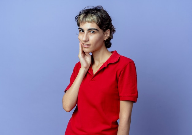 Thoughtful young caucasian girl with pixie haircut putting hand on cheek isolated on purple background with copy space