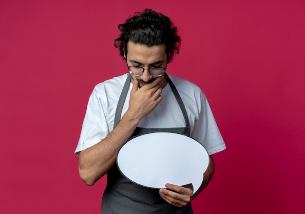 Thoughtful young caucasian male barber wearing glasses and wavy hair band in uniform holding chat bubble putting hand on chin looking down isolated on crimson background with copy space