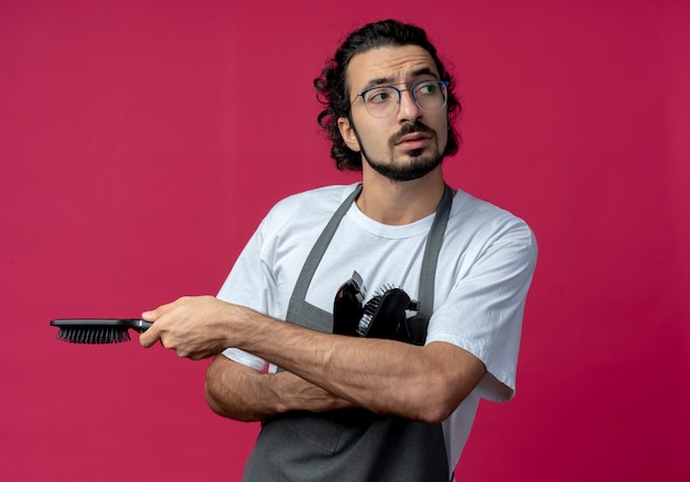 Free photo thoughtful young caucasian male barber wearing glasses and wavy hair band in uniform holding hair clippers, comb, spray bottle looking at side isolated on crimson background