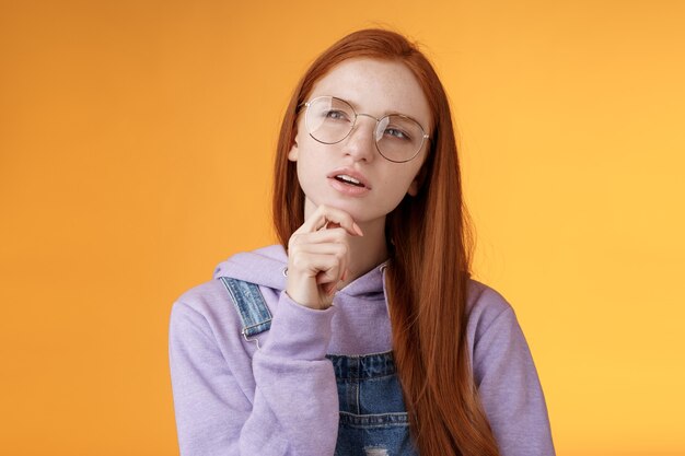 Thoughtful young creative smart redhead girl thinking figure out important thing standing upper left corner squinting thinking get clue touch chin thinking, pondering choice, orange background