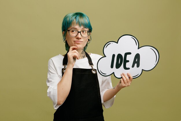 Thoughtful young female barber wearing glasses uniform looking at side keeping hand on chin showing idea bubble isolated on olive green background