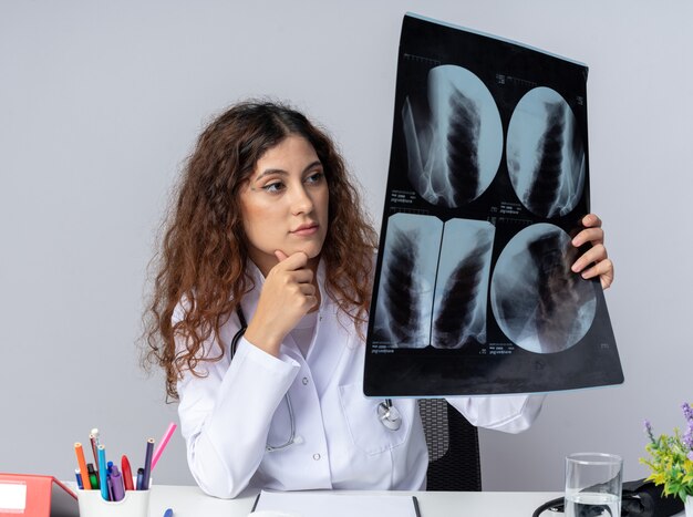 Thoughtful young female doctor wearing medical robe and stethoscope sitting at table with medical tools keeping hand on chin holding and looking at x-ray shot isolated on white wall