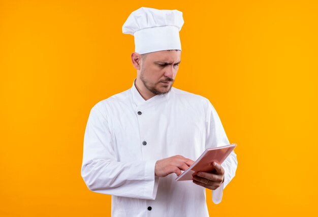 Thoughtful young handsome cook in chef uniform holding note pad putting hand on and looking at it isolated on orange space