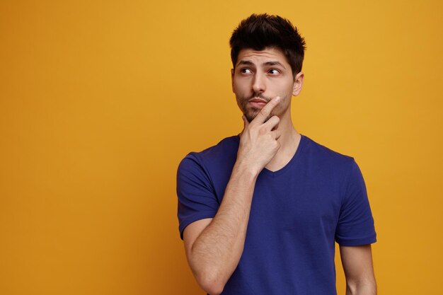 Thoughtful young handsome man keeping hand on chin looking at side on yellow background with copy space