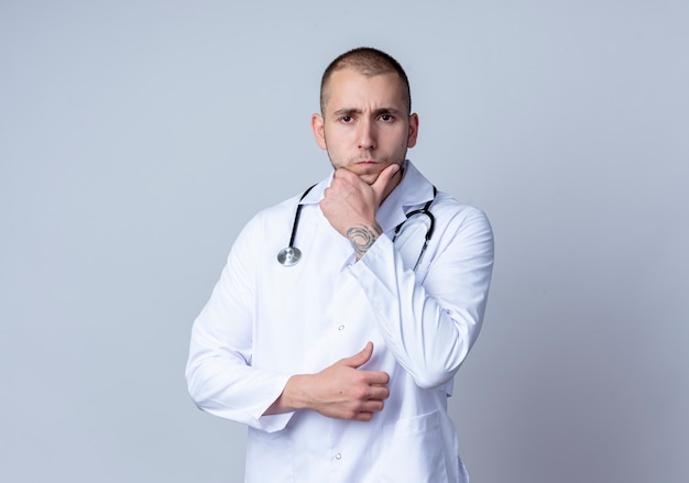 Thoughtful young male doctor wearing medical robe and stethoscope around his neck putting hand on chin isolated on white wall