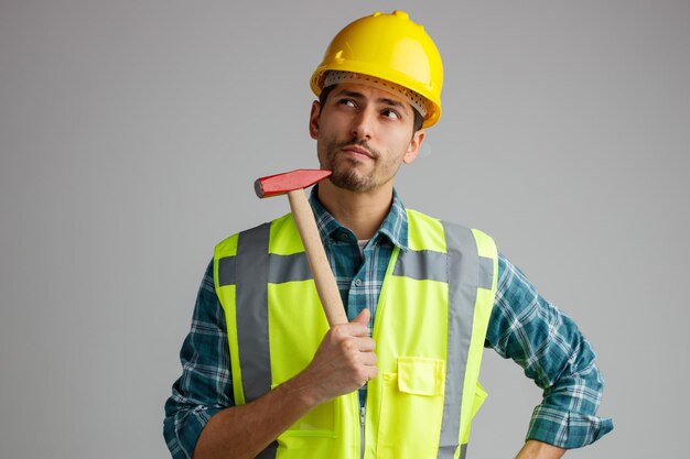 Thoughtful young male engineer wearing safety helmet and uniform touching chin with hammer looking up keeping hand on waist isolated on white background