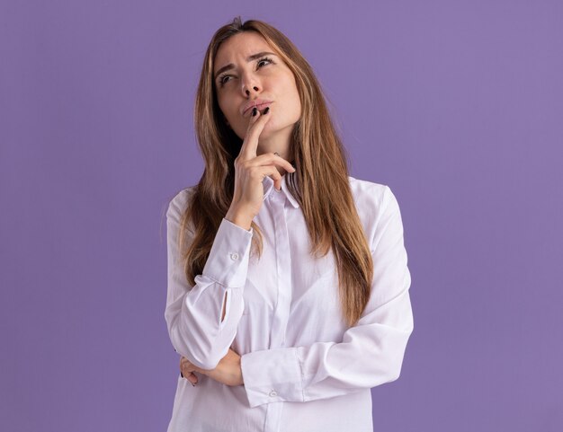 Thoughtful young pretty caucasian girl puts hand on chin and looks up on purple 