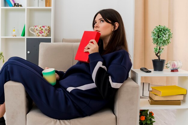 Thoughtful young pretty caucasian woman sitting on armchair in designed living room holding book and cup looking at side and touching chin with book