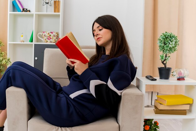 Thoughtful young pretty caucasian woman sitting on armchair in designed living room reading book