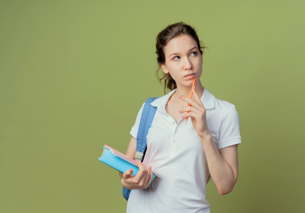 Thoughtful young pretty female student wearing back bag looking at side holding note pad and book and touching chin with pen isolated on olive green background with copy space