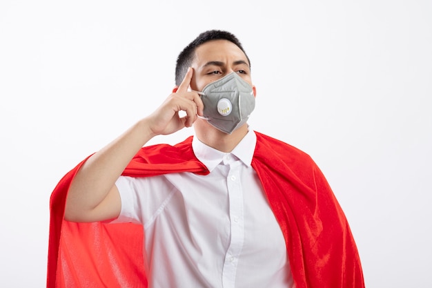 Free photo thoughtful young superhero boy in red cape wearing protective mask looking at side doing think gesture isolated on white background