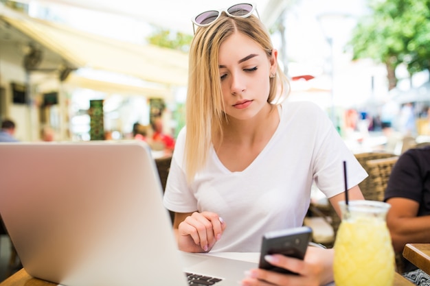 Thoughtful young woman holds phone, uses laptop computer for online communication