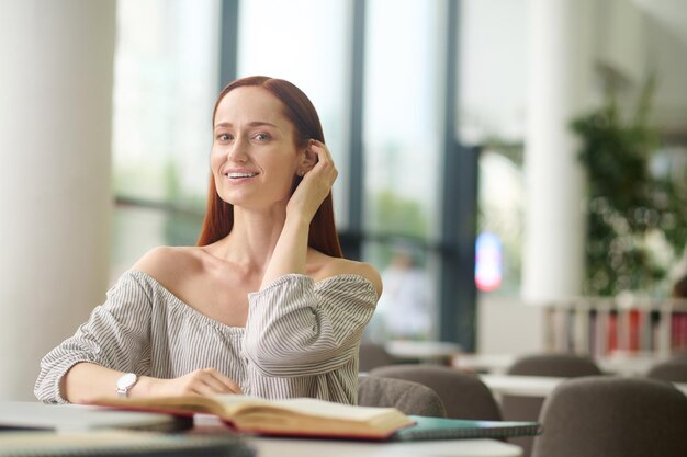 Thoughts, idea. Young smiling woman touching long red hair sitting at table with books thinking in good mood in room during day