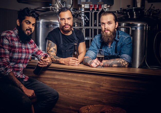 Three bearded interracial friends drink craft beer in a brewery. Two hipster workers in aprons drink beer with their friend in a brewery factory.