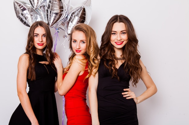 Three best friends celebrate birthday indoor wearing elegant evening dresses and have bright makeup. Girls hugging and Showing signs with their hands.