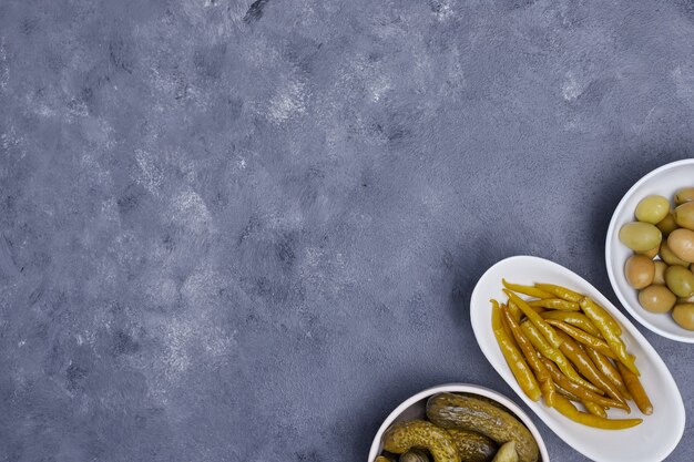 Three bowls of pickled cucumbers, peppers and olives on blue background.