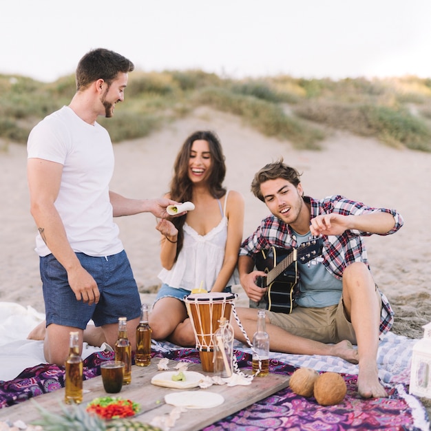 Three friends sitting at the beach with guitar and drinks