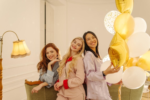 Three gorgeous young interracial girls in silk pajamas are celebrating birthday in room with balloons Concept of playful mood holiday