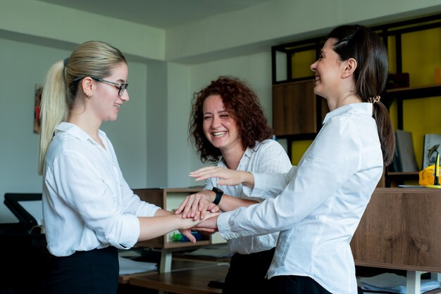 Three happy satisfied women office coworkers smiling cheerfully stacked their hands together gesture of friendship unity and partnership in business standing in office