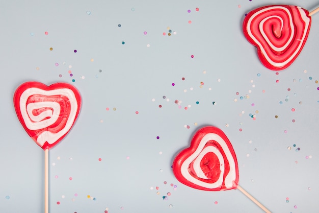 Free Photo three heart shape red lollipops on gray background with colorful confetti