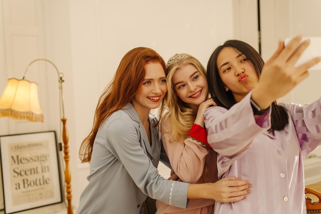 Three interracial young girls in pajamas take photo together on smartphone indoors Entertainment and lifestyle concept