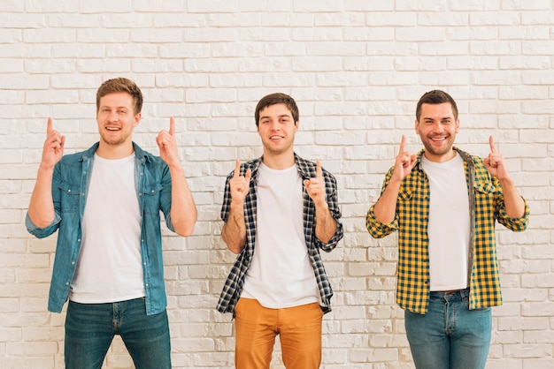 Three male friends standing against white wall pointing finger upward looking at camera