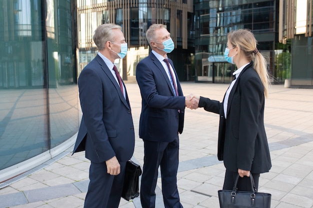 Free photo three office workers in facial masks making deal or greeting. professional successful businesswoman and businessmen standing outdoors and handshaking. negotiation, protection and partnership concept