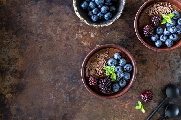 Three ramekins with chocolate mousse with blueberries blackberries and fresh mint on a rusty iron background