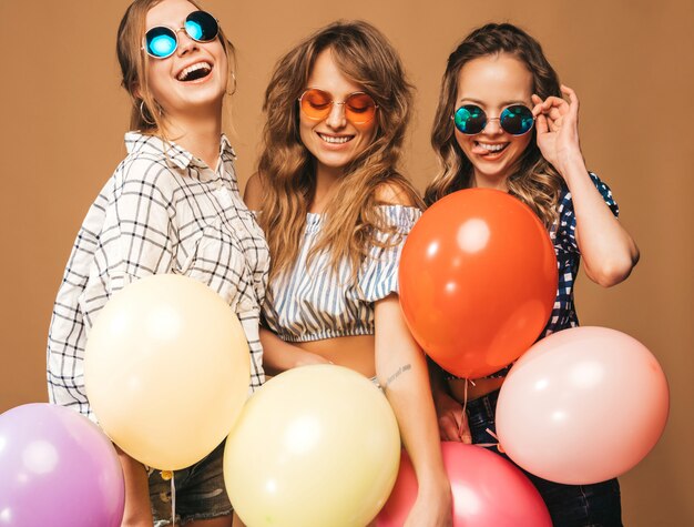 Three smiling beautiful women in checkered shirt summer clothes and sunglasses. Girls posing. Models with colorful balloons. Having fun, ready for celebration birthday 