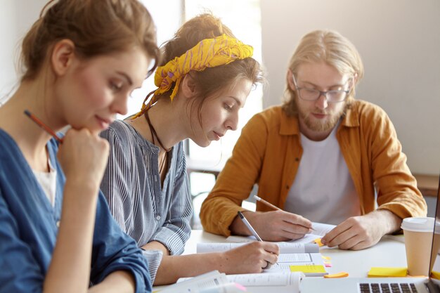 Three students sitting together at their working place, writing with pencils and studying scientific literature, preparing for exams in university. Bearded guy and two females working on project