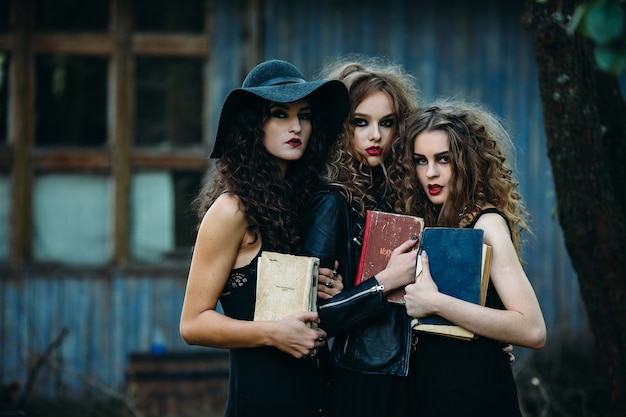 Three vintage women as witches, pose in front of an abandoned building with books in hand on the eve of Halloween