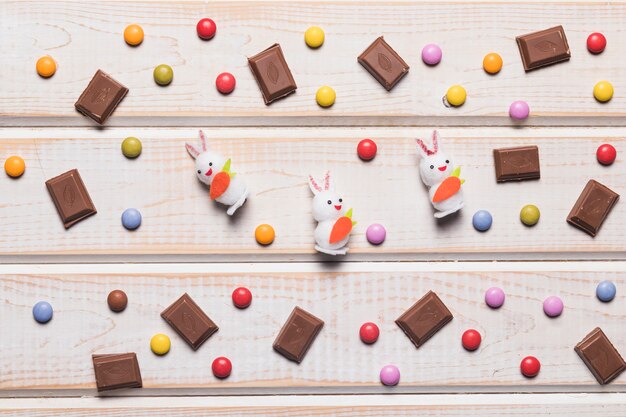 Three white bunnies surrounded with multicolored gems and chocolate pieces over desk