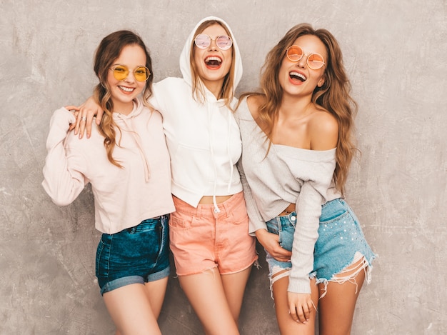 Three young beautiful smiling girls in trendy summer sport clothes. Sexy carefree women posing. Positive models in round sunglasses having fun. Hugging