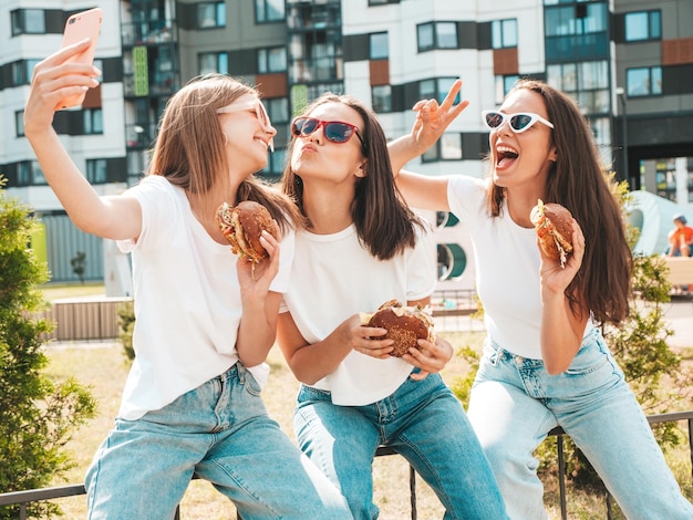 Free photo three young beautiful smiling hipster female in trendy summer clothessexy carefree women posing in the streetpositive models taking selfie in sunglassesholding juicy burger and eating hamburger