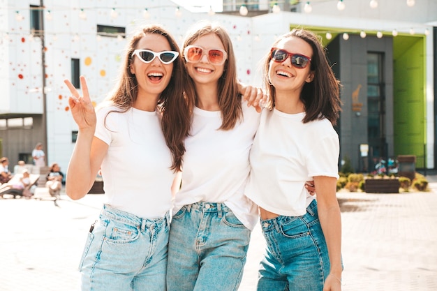 Three young beautiful smiling hipster female in trendy summer same clothes Sexy carefree women posing on the street backgroundPositive models having fun in sunglassesHugging Cheerful and happy