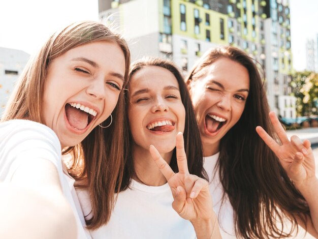 Three young beautiful smiling hipster female in trendy summer same clothes Sexy carefree women posing on the street backgroundPositive models having fun in sunglassesTaking Pov selfie photos