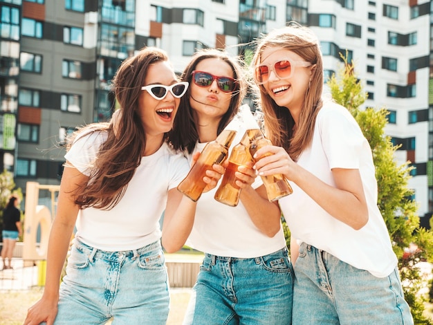 Three young beautiful smiling hipster female in trendy summer same clothesSexy carefree women posing in the streetPositive models having fun in sunglasses Drinking bottle beer Oktoberfest