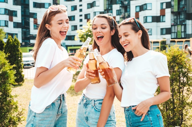 Three young beautiful smiling hipster female in trendy summer same clothesSexy carefree women posing in the streetPositive models having fun in sunglasses Drinking bottle beer Oktoberfest