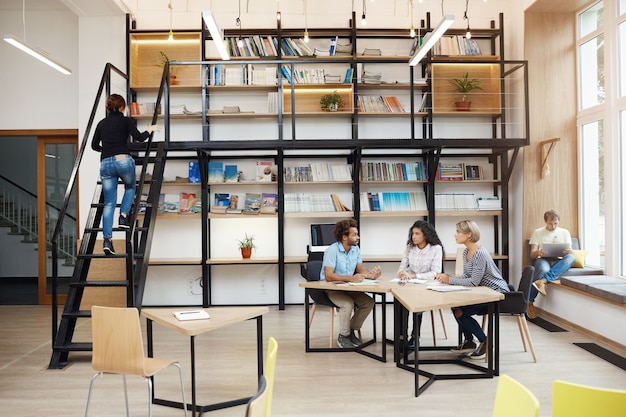 Free Photo three young perspective startupers sitting in modern light library on meeting, talking about new project, looking though details of work, having productive day in friends atmosphere,