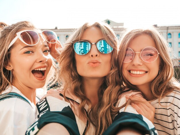 Three young smiling hipster women in summer clothes.Girls taking selfie self portrait photos on smartphone.Models posing in the street.Female showing positive face emotions in sunglasses