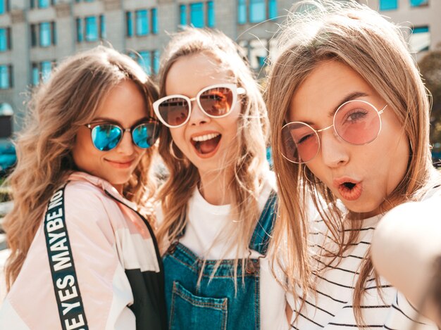 Three young smiling hipster women in summer clothes.Girls taking selfie self portrait photos on smartphone.Models posing in the street.Female showing positive face emotions