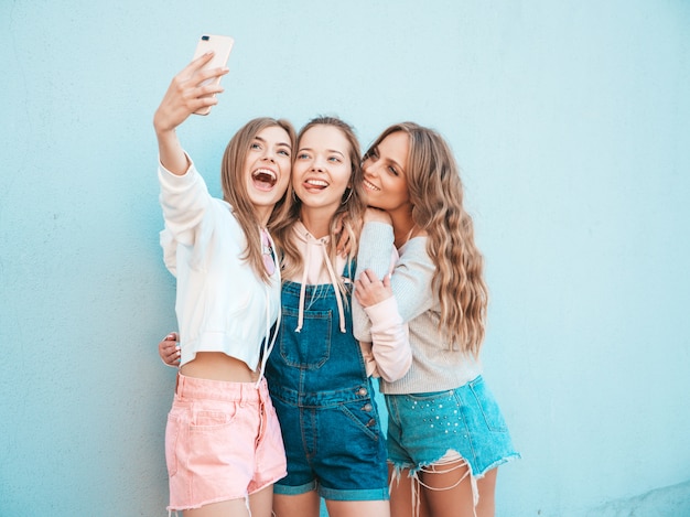 Three young smiling hipster women in summer clothes.Girls taking selfie self portrait photos on smartphone.Models posing in the street near wall.Female showing positive face emotions.Showing tongue