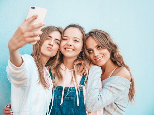 Three young smiling hipster women in summer clothes.Girls taking selfie self portrait photos on smartphone.Models posing in the street near wall.Female showing positive face emotions