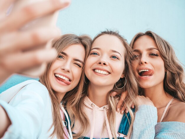 Three young smiling hipster women in summer clothes.Girls taking selfie self portrait photos on smartphone.Models posing in the street near wall.Female showing positive face emotions.