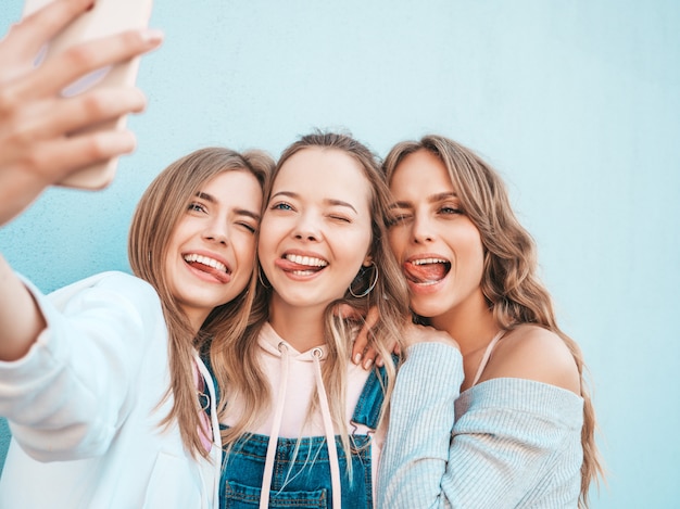 Free photo three young smiling hipster women in summer clothes.girls taking selfie self portrait photos on smartphone.models posing in the street near wall.female showing positive face emotions