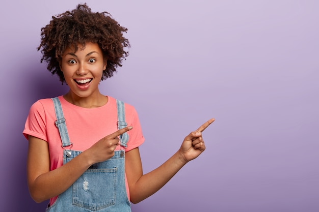 Thrilled cheerful Afro American curly girl gives way to awesome place, points aside, smiles broadly, looks happily at camera, gestures against purple background