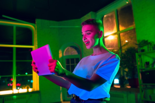 Thrilled. Cinematic portrait of stylish woman in neon lighted interior. Toned like cinema effects, bright neoned colors. Caucasian model using tablet in colorful lights indoors. Youth culture.